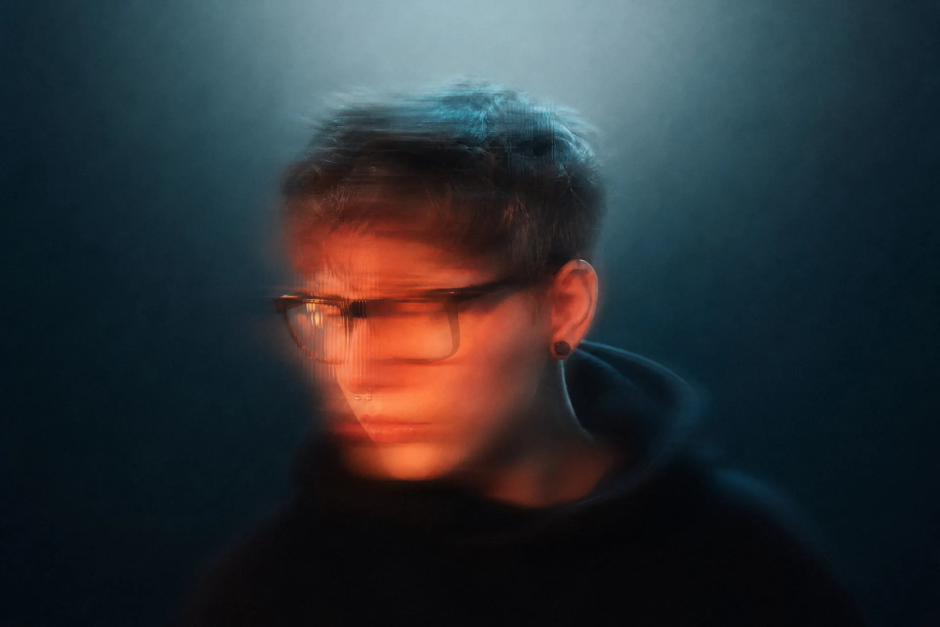 Portrait of OnlyRodas in motion, wearing glasses and a black hoodie, lit in orange against a dark teal background